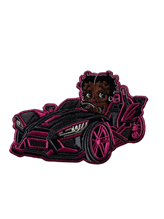 An embroidered patch featuring a cartoon-style Black woman with braided hair driving a pink and black sports car, shown on a light textured background.