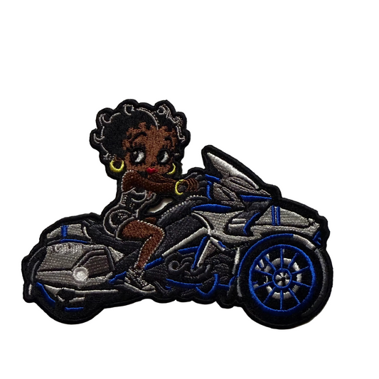 An embroidered patch featuring a stylized cartoon woman with dark curly hair riding a silver and blue motorcycle, shown against a plain off-white background.